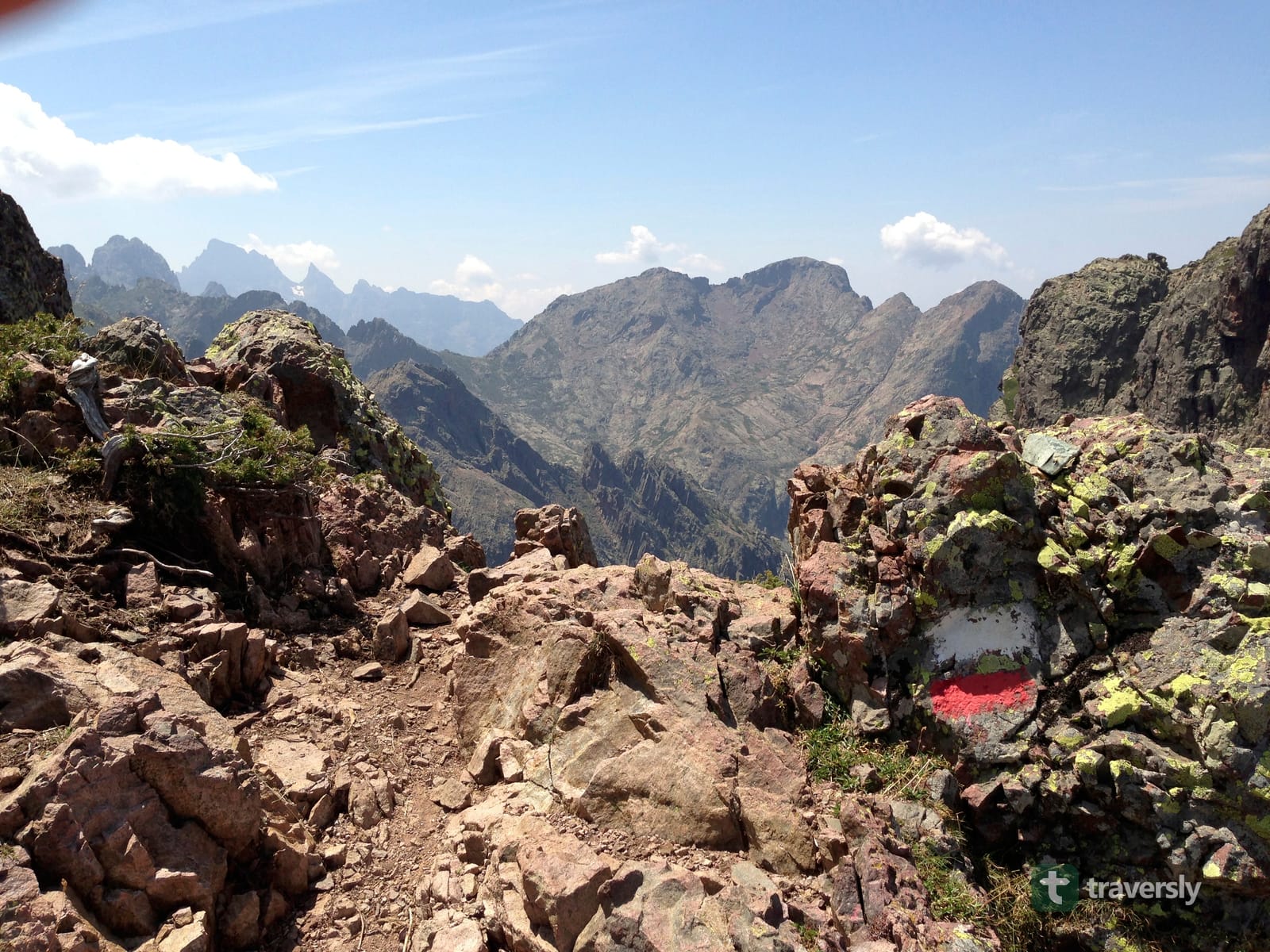 A red-and-white GR20 waymark painted on a granite outcrop, with Corsica's interior peaks behind.