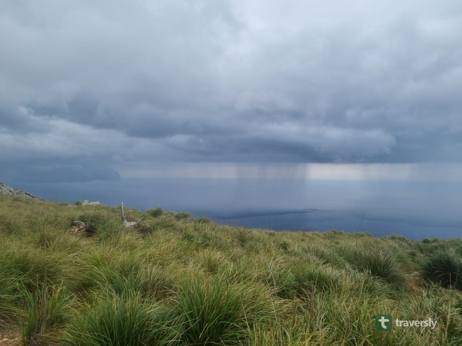 A dark rain shaft falls from storm clouds over the Mediterranean, seen from a ridge on the GR221.