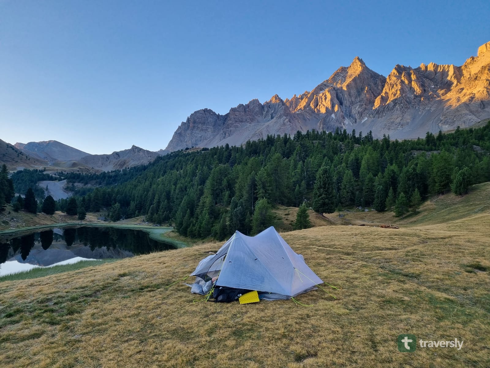 Sunrise alpenglow on the peaks above a mountain tarn on the GR5 in the French Alps, a lightweight tent pitched on the grass in the foreground.