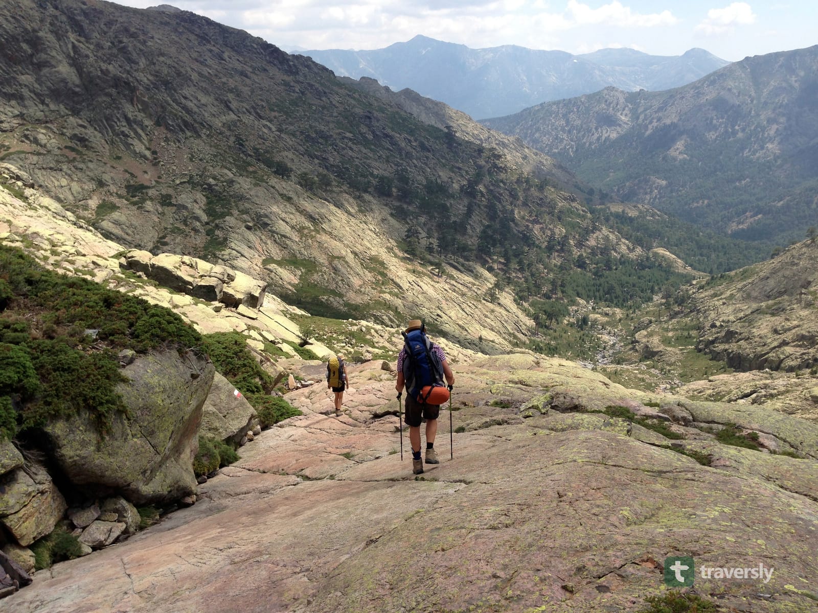 Two hikers with backpacks descending a bare granite slope on the GR20 in Corsica, mountains and a valley below.